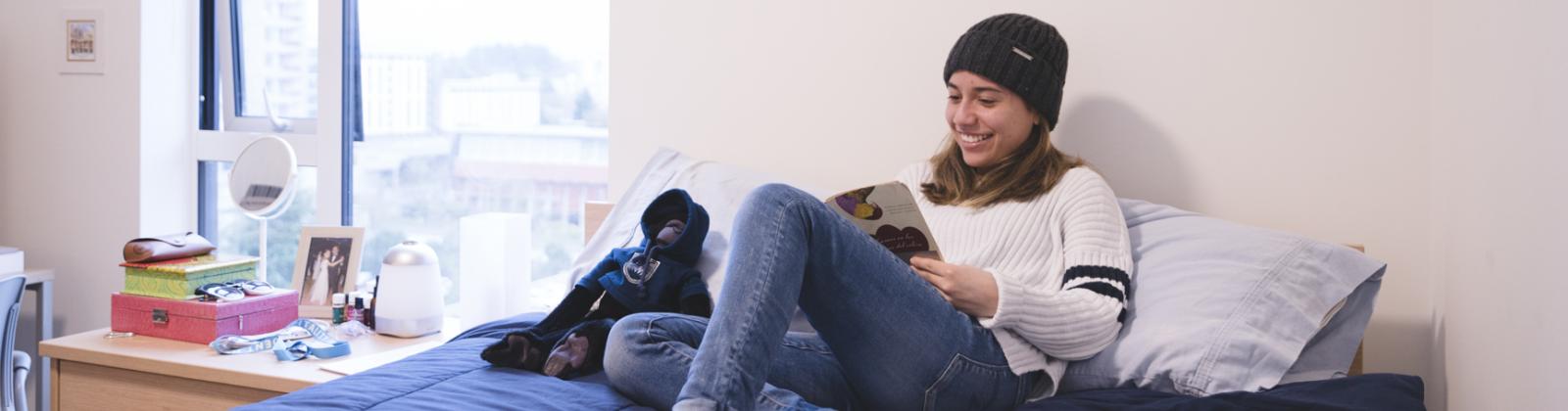 A student smiling and laying upright in her residence room bed while reading a book