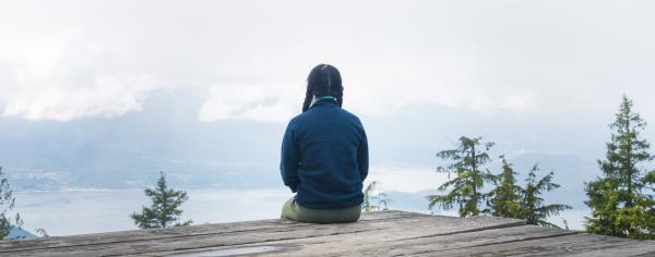 A photograph of the back of a person sitting on a wooden ledge overlooking the water and mountains of BC.