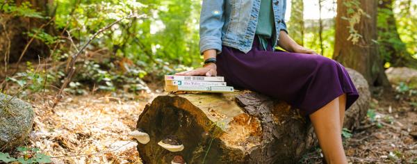 Student sitting on a stump in the forest while touching a stack of books
