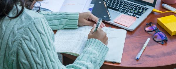 A student wearing a mint-coloured sweater sitting at a desk and writing in a notebook with her laptop in front of her
