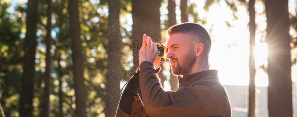 A male student wearing a brown jacket standing in a forested area while looking through a small yellow device that he's holding up to his eye