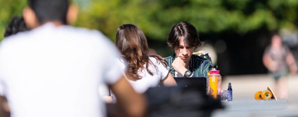 A student sitting outdoors at a picnic table working on their laptop while in deep thought 