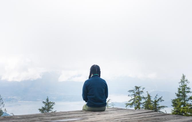 A photograph of the back of a person sitting on a wooden ledge overlooking the water and mountains of BC.