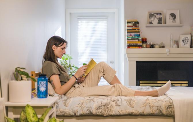 A photograph of a student sitting on their bed reading a book. Their back is against the wall and their legs are stretched forward. There is a nightstand in the foreground and a window and fireplace in the background.