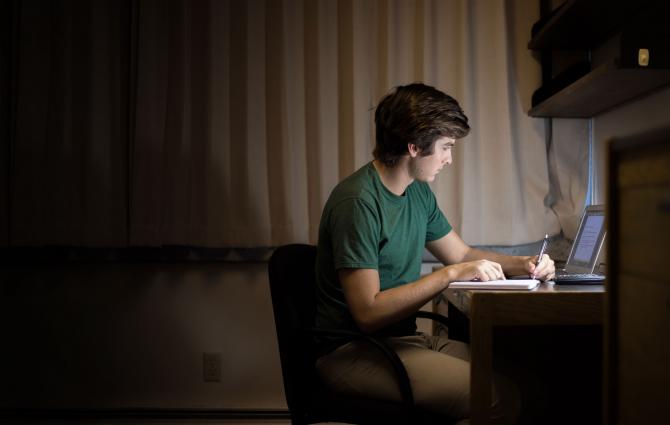 A student studying at his desk during the evening, his face illuminated by his laptop screen