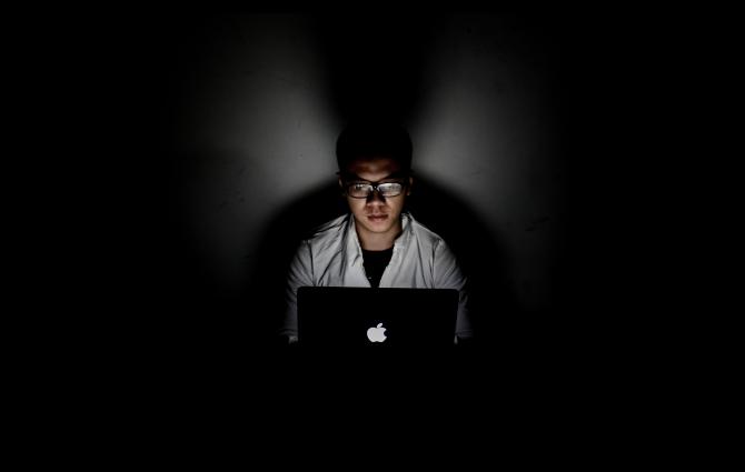 A student studying in a dark room, his face illuminated by the laptop screen