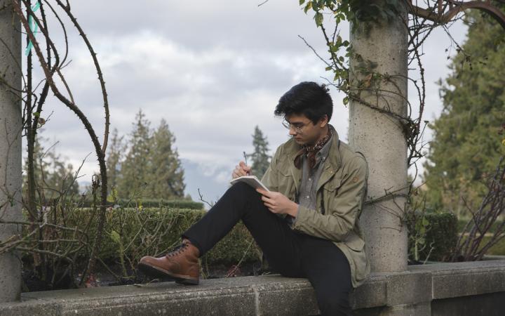 A photograph of a student sitting on a ledge in the Rose Garden at UBC, journalling. 