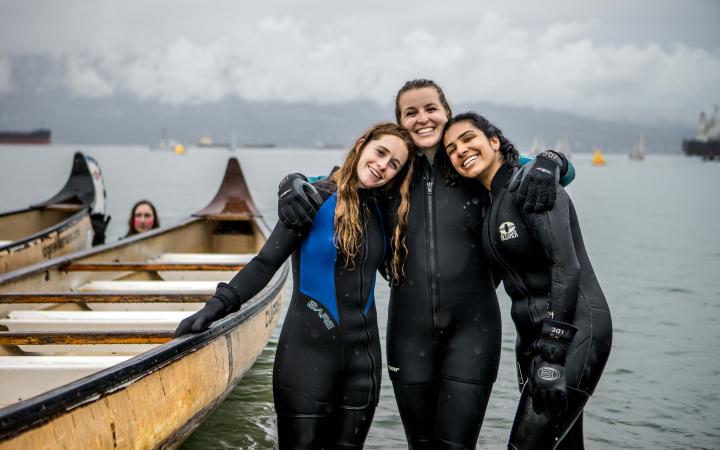 Three students in wetsuits hugging next to a longboat