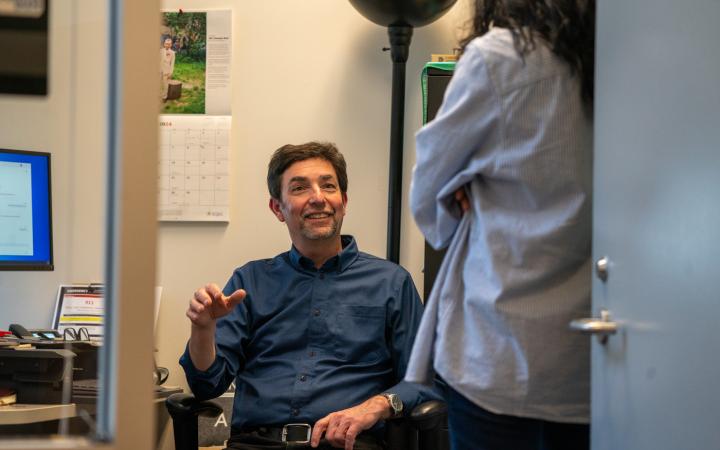 Tony Botelho sitting in a chair in his office while looking up at someone, smiling and engaging in conversation
