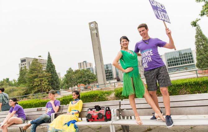 A pair of dancers standing next to a boom box on a bench overlooking the UBC clock tower and Irving K Barber Learning Centre.